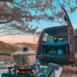 A camping setup with a gas stove and pot on a table in the foreground, and an open SUV with a sleeping area in the back, set outdoors at sunset.