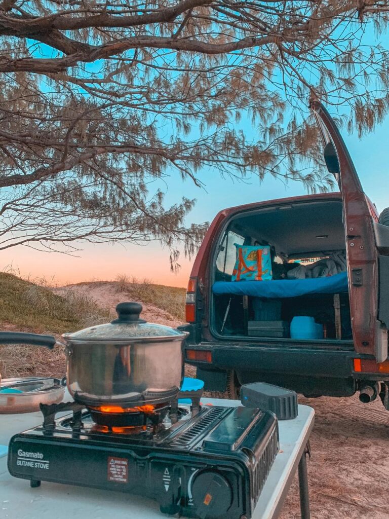 A camping setup with a gas stove and pot on a table in the foreground, and an open SUV with a sleeping area in the back, set outdoors at sunset.
