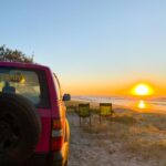 A red SUV is parked on sandy ground near the beach at sunset, with two empty camping chairs facing the ocean.