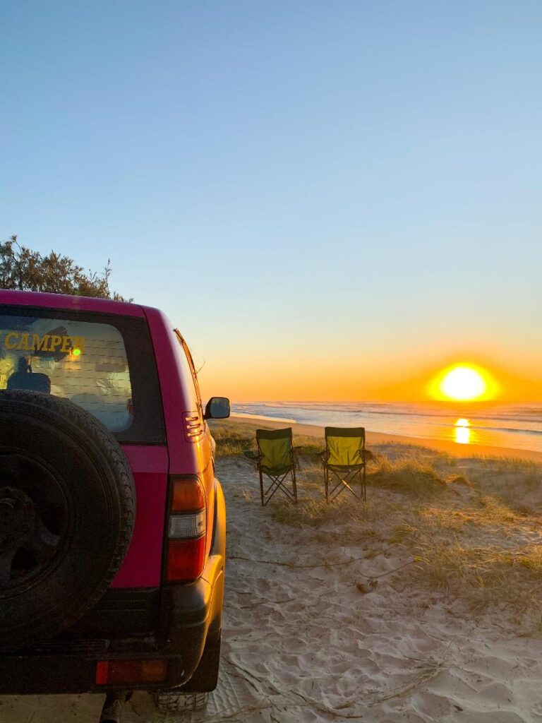 A red SUV is parked on sandy ground near the beach at sunset, with two empty camping chairs facing the ocean.