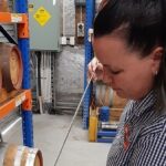 A person in a checked shirt draws liquid from a wooden barrel with a sampling tool in a warehouse filled with stacked barrels.