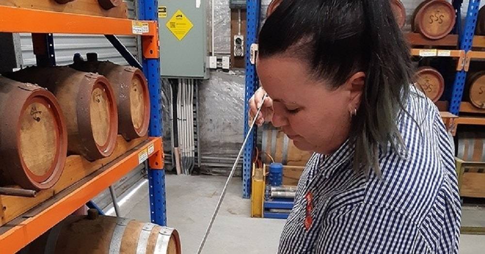 A person in a checked shirt draws liquid from a wooden barrel with a sampling tool in a warehouse filled with stacked barrels.