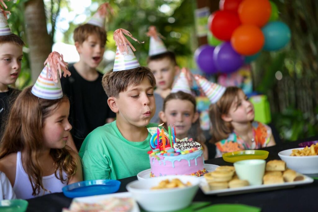 A boy wearing a party hat blows out candles on a birthday cake surrounded by other children at an outdoor party with balloons and snacks.