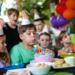 A boy wearing a party hat blows out candles on a birthday cake surrounded by other children at an outdoor party with balloons and snacks.