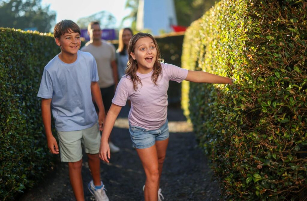 Two children smiling and walking through a hedge maze, followed by two adults in the background on a sunny day.