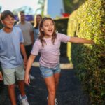 Two children smiling and walking through a hedge maze, followed by two adults in the background on a sunny day.