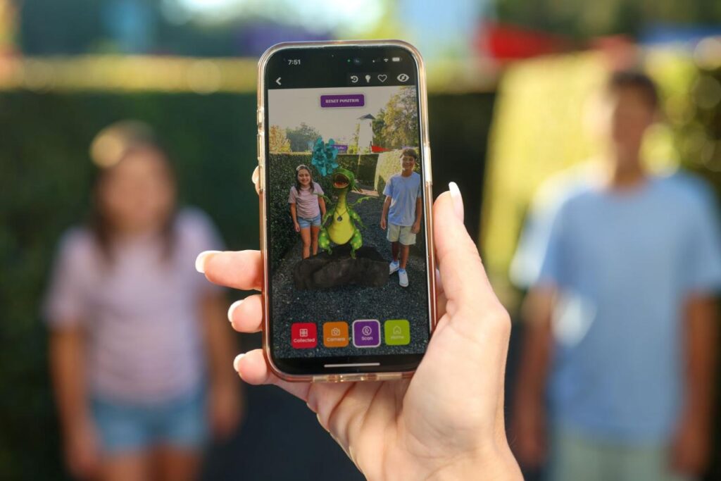 A person holds a smartphone showing an augmented reality image of two people standing beside a cartoonish green creature, while two people stand blurred in the background.