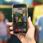 A person holds a smartphone showing an augmented reality image of two people standing beside a cartoonish green creature, while two people stand blurred in the background.