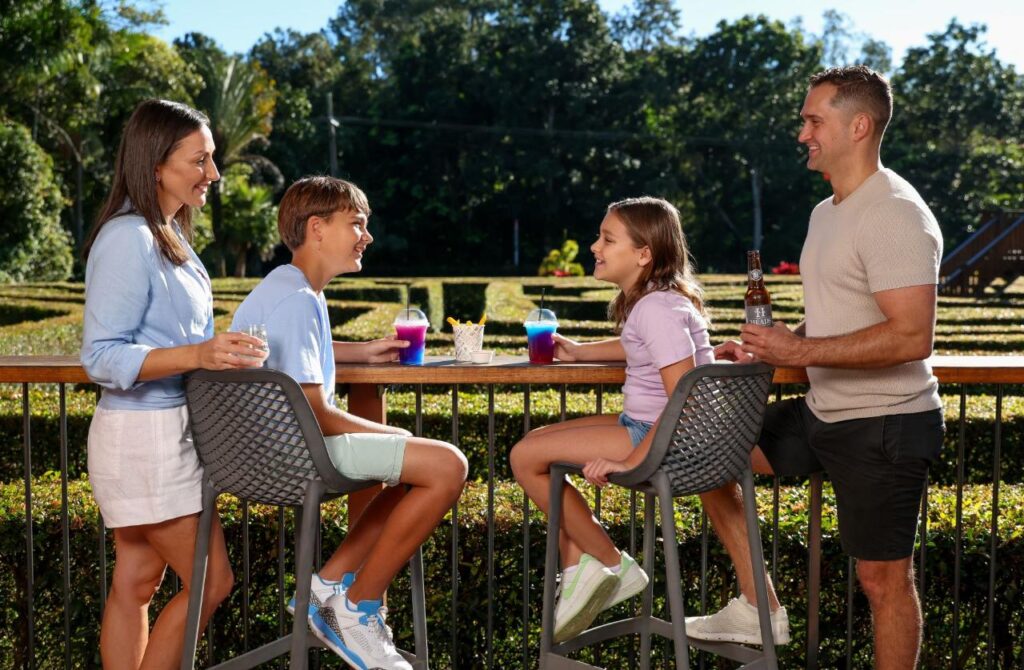 A family of four sits and stands around an outdoor bar table, holding drinks and smiling, with a garden and trees in the background.