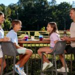 A family of four sits and stands around an outdoor bar table, holding drinks and smiling, with a garden and trees in the background.