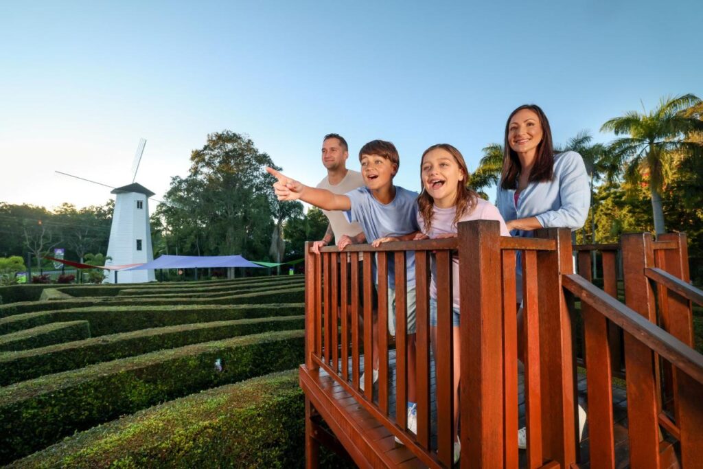A family of four stands on a wooden platform overlooking a hedge maze, with a white windmill and trees in the background. One child excitedly points ahead.