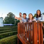 A family of four stands on a wooden platform overlooking a hedge maze, with a white windmill and trees in the background. One child excitedly points ahead.