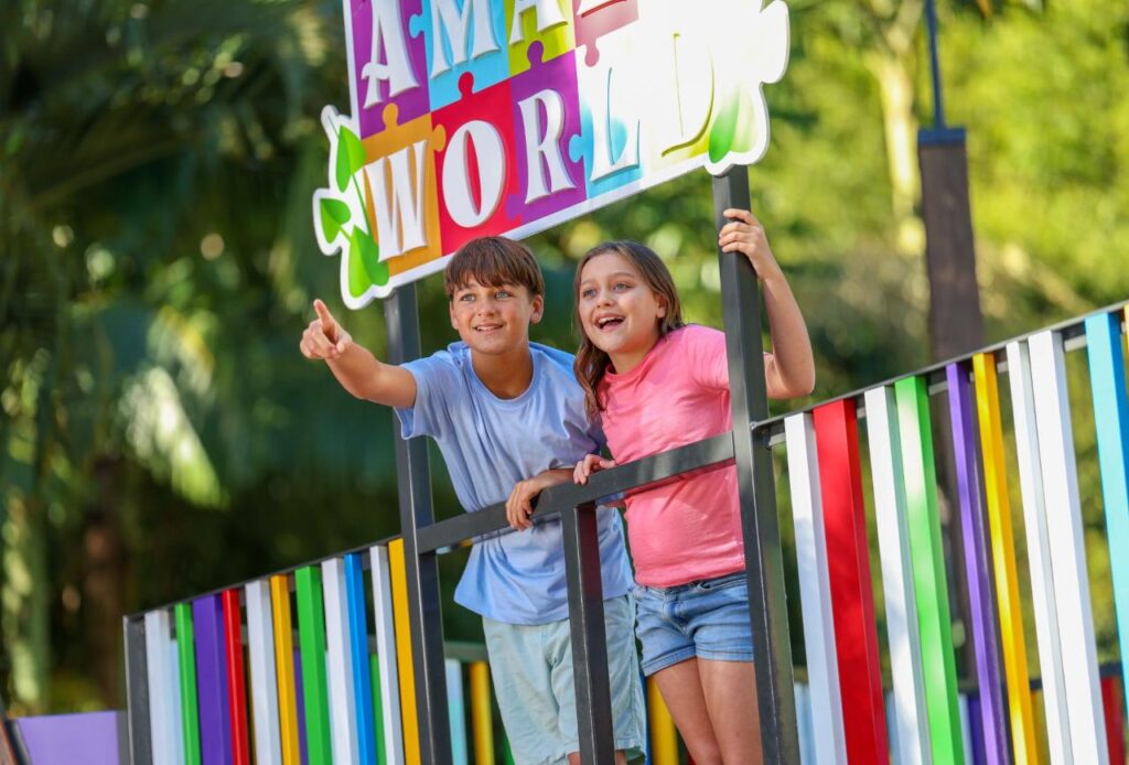 Two children stand on a colorful platform, smiling and pointing, under a sign that reads "AMAZE WORLD" with trees in the background.