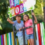 Two children stand on a colorful platform, smiling and pointing, under a sign that reads "AMAZE WORLD" with trees in the background.