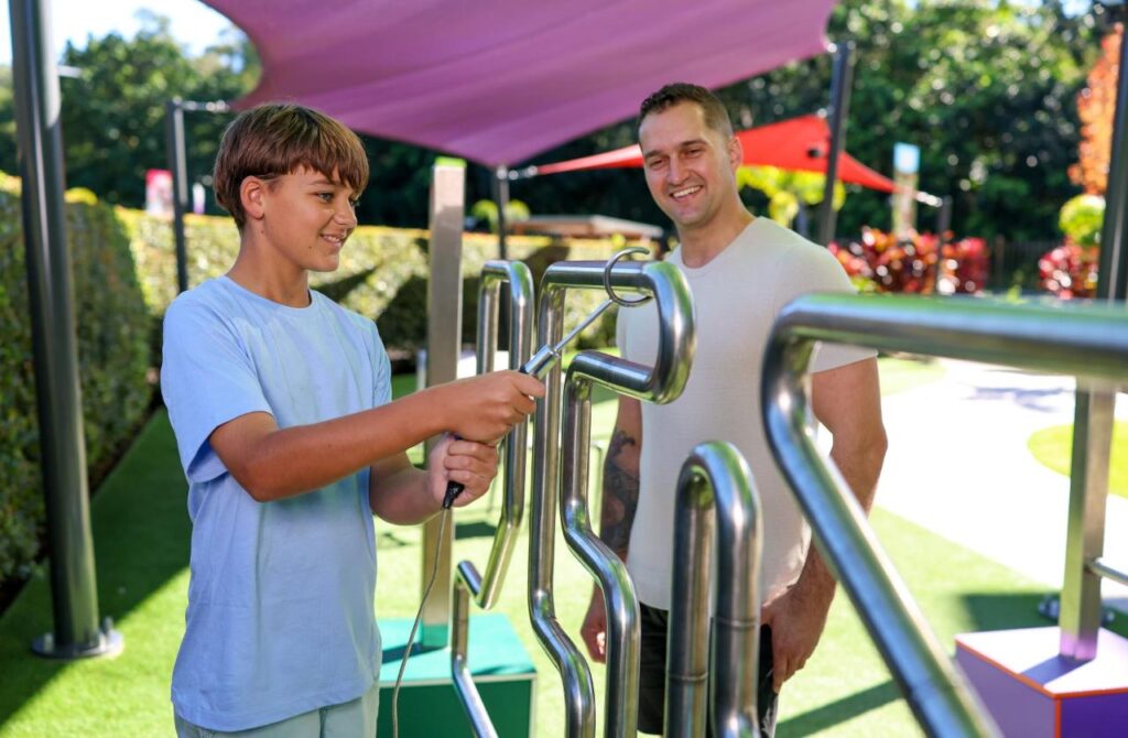 A boy in a light blue shirt uses a metal hand maze outdoors while an adult man watches and smiles. The setting is a sunny, colorful playground.