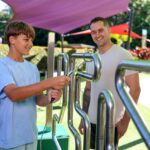 A boy in a light blue shirt uses a metal hand maze outdoors while an adult man watches and smiles. The setting is a sunny, colorful playground.