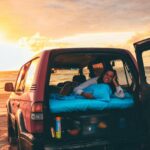 A person lies on a mattress in the back of an SUV parked on a sandy beach at sunset, with the rear door open and various items nearby.