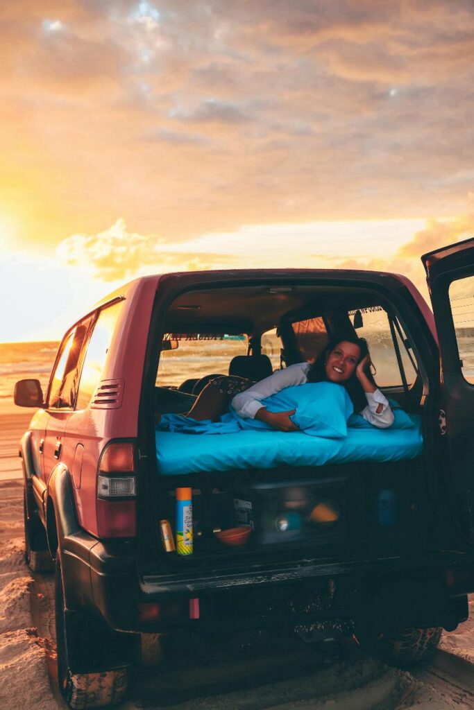 A person lies on a mattress in the back of an SUV parked on a sandy beach at sunset, with the rear door open and various items nearby.