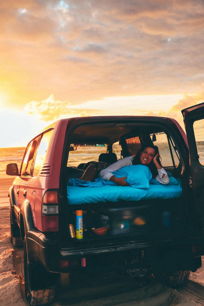 A person lies on a mattress in the back of an SUV parked on a sandy beach at sunset, with the rear door open and various items nearby.