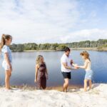 A man helps a young girl climb a sandy bank near a lake, while another woman and girl look on under a partly cloudy sky.