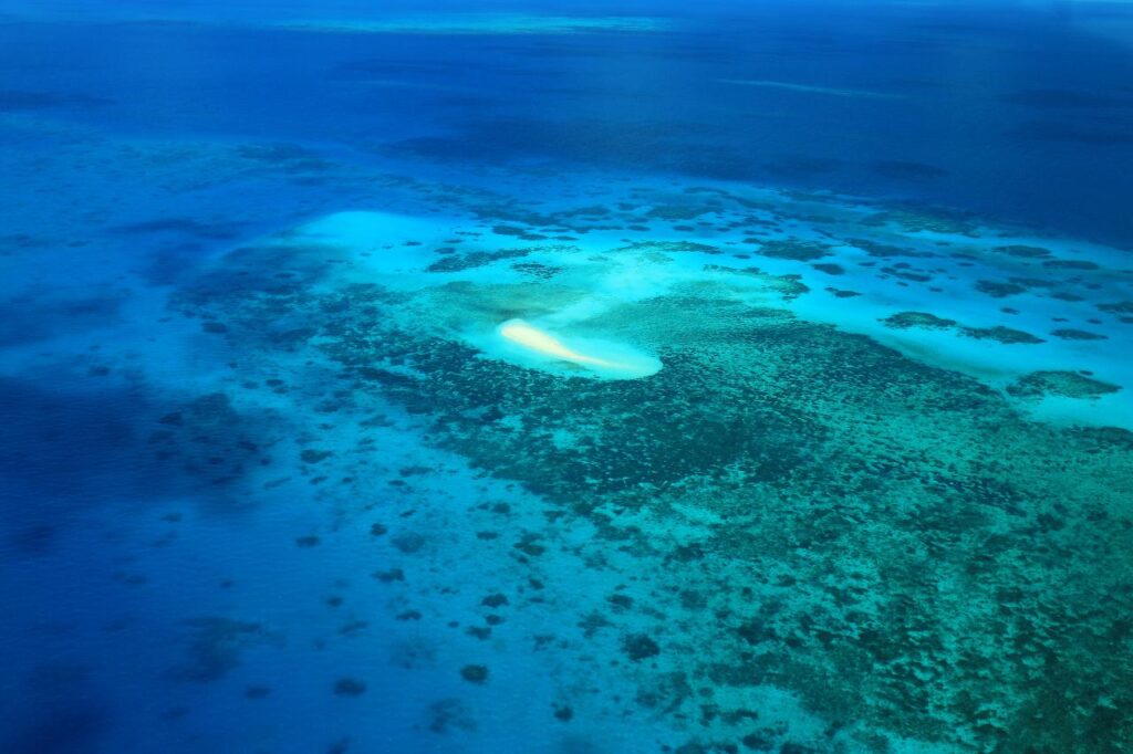 Aerial view of a small sand island surrounded by shallow turquoise water and coral reefs in the ocean.