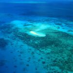 Aerial view of a small sand island surrounded by shallow turquoise water and coral reefs in the ocean.