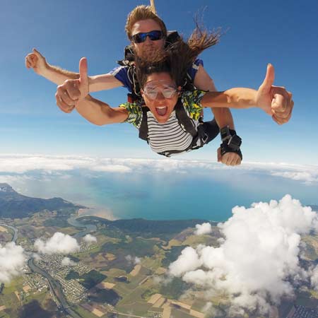 Two people tandem skydiving above a landscape with clouds, fields, and water visible below; both are smiling and giving thumbs-up gestures.