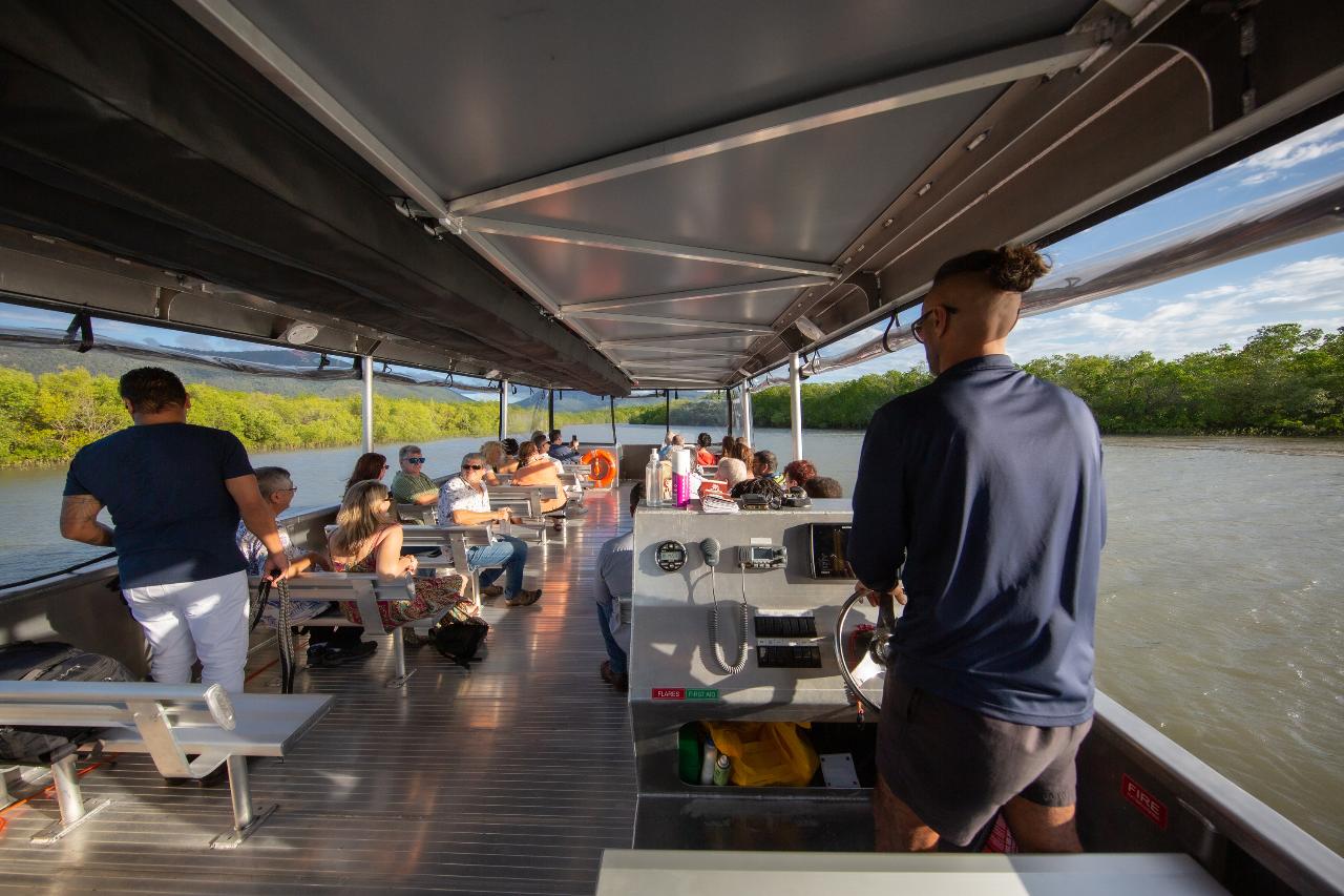 Passengers sit and stand on a covered riverboat as it travels along a river surrounded by greenery under a partly cloudy sky.