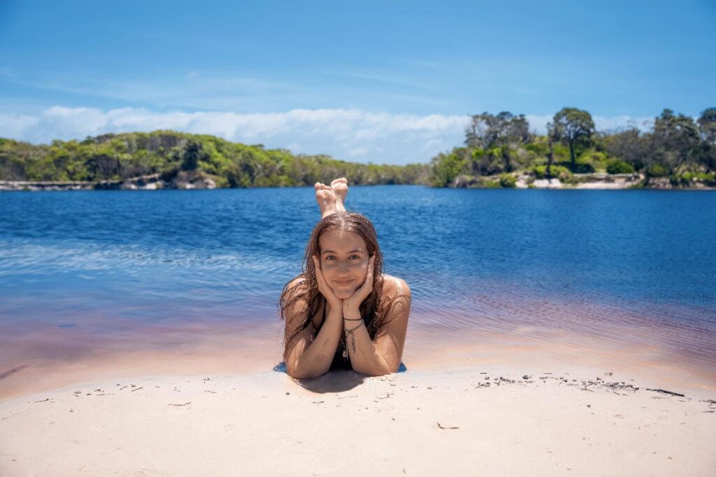 A young woman lies on a sandy shore with her chin in her hands, smiling, in front of a blue lake surrounded by trees under a clear sky.