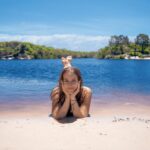 A young woman lies on a sandy shore with her chin in her hands, smiling, in front of a blue lake surrounded by trees under a clear sky.