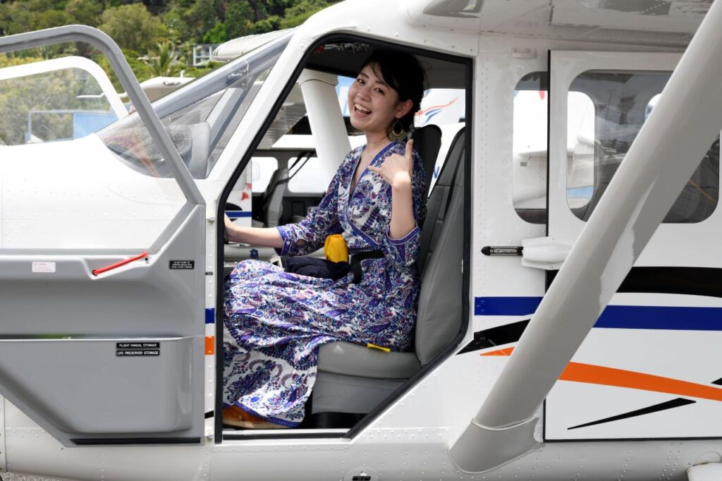 A woman in a blue patterned dress sits in the pilot seat of a small airplane, smiling and gesturing with her hand, with the plane door open.