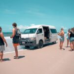 A group of people stands on a sandy beach near a white van stuck close to the shoreline under a clear sky.
