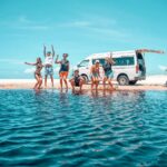 Six people in swimwear pose and jump on a sandy beach near a blue van with an open door, with clear sky and blue water in the foreground.