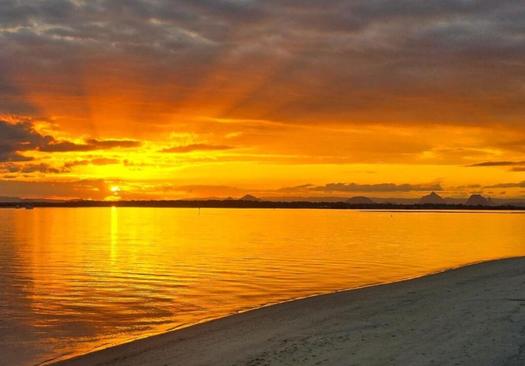 Bright orange and yellow sunset over calm water, with sun rays visible through clouds and a sandy shore in the foreground.