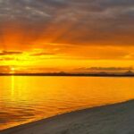Bright orange and yellow sunset over calm water, with sun rays visible through clouds and a sandy shore in the foreground.