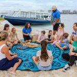 A group of adults and children sit in a circle on a patterned blue mat on a sandy beach near a docked boat, listening to a woman speak.