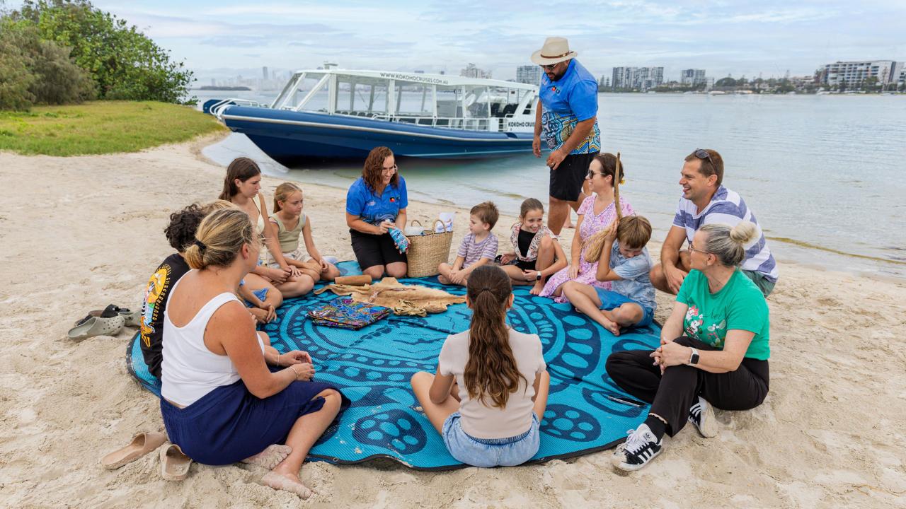A group of adults and children sit in a circle on a patterned blue mat on a sandy beach near a docked boat, listening to a woman speak.