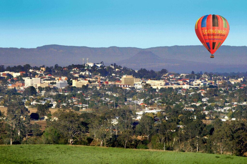 A red and orange hot air balloon floats above a town with scattered buildings, trees, and distant hills under a clear blue sky.