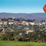 A red and orange hot air balloon floats above a town with scattered buildings, trees, and distant hills under a clear blue sky.