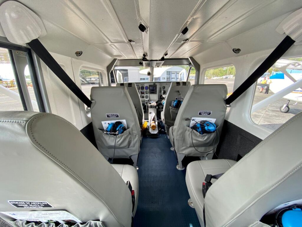 Interior view of a small airplane showing four seats, seat belts, headsets, and the cockpit controls at the front.