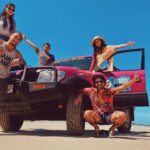 Five people pose around a pink SUV parked on a sandy beach under a clear blue sky; two stand on the vehicle while others pose in front and beside it.