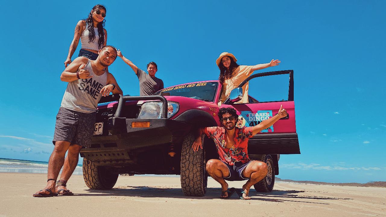 Five people pose around a pink SUV parked on a sandy beach under a clear blue sky; two stand on the vehicle while others pose in front and beside it.