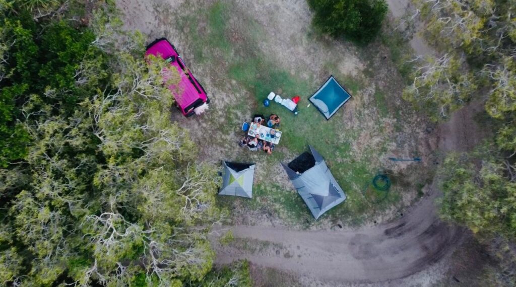 Aerial view of a campsite with three tents, a picnic table with people seated, a pink vehicle, and surrounding trees on a grassy area near a dirt road.