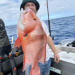 A person on a boat holds a large red fish, partially obscuring their face, with the ocean and a fishing rod in the background.
