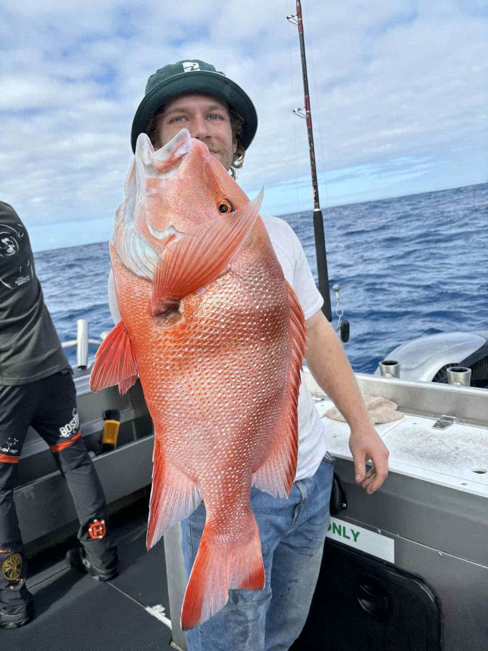 A person on a boat holds a large red fish, partially obscuring their face, with the ocean and a fishing rod in the background.
