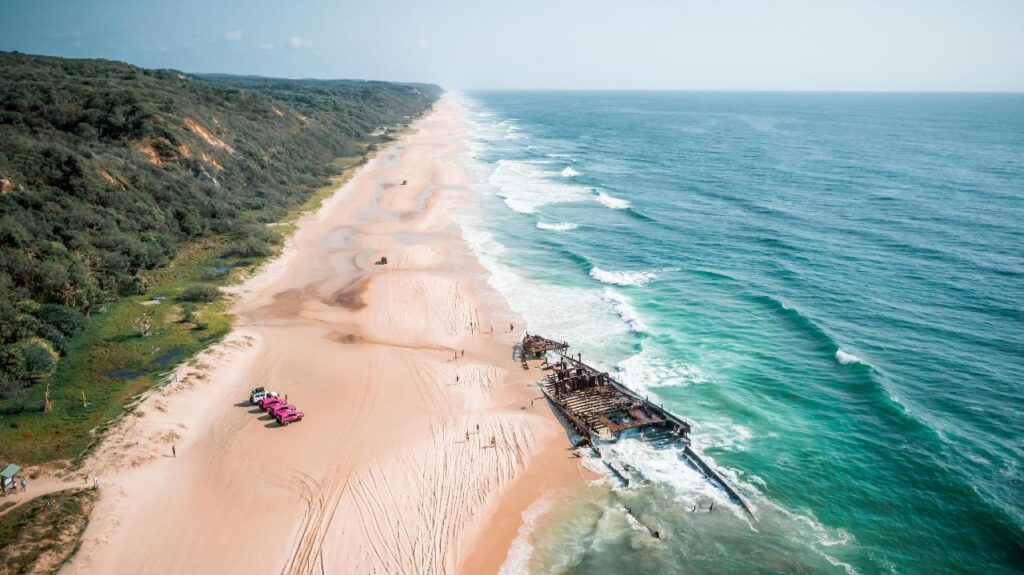 Aerial view of a long sandy beach with waves, a rusty shipwreck near the shore, and several pink vehicles parked on the sand beside a green, forested area.