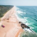 Aerial view of a long sandy beach with waves, a rusty shipwreck near the shore, and several pink vehicles parked on the sand beside a green, forested area.