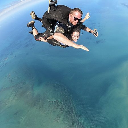 Two people tandem skydiving over a large body of water, with the Earth visible far below and clear skies above.