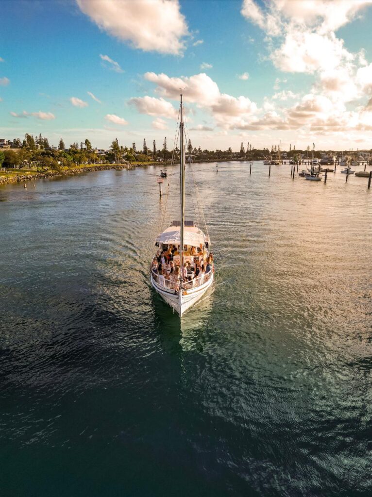 A sailboat with people on board moves through calm water near a marina, with a partly cloudy sky and trees visible in the background.