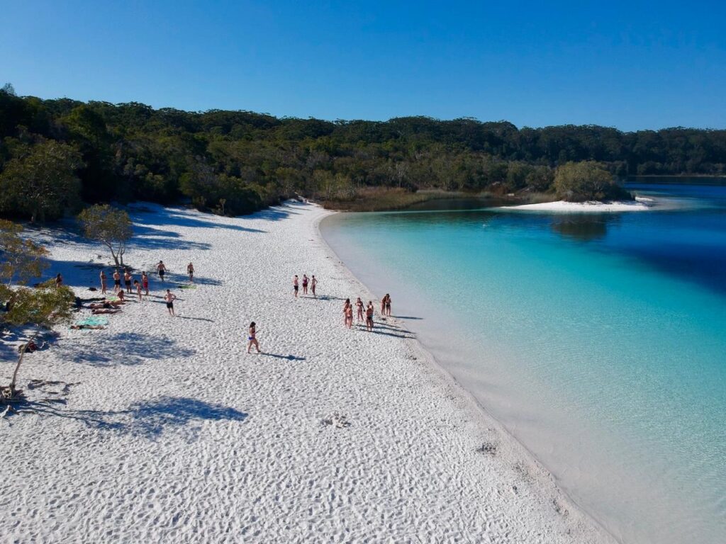 A group of people walk and relax on a white sandy beach beside clear blue water, with forested land in the background under a clear sky.